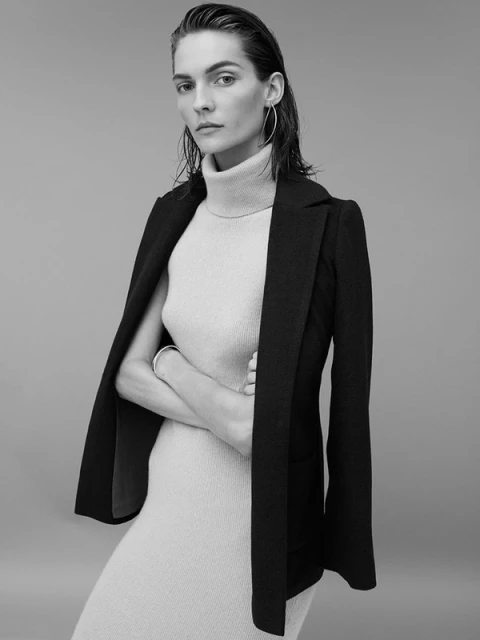 Monochrome portrait of an adult woman in a turtleneck dress, layered with a blazer, posing indoors with her arms crossed.
