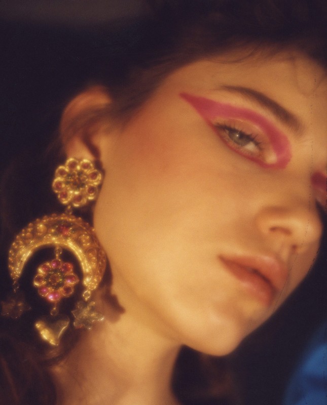 Close-up portrait of a woman with pink eye makeup, dramatic gold earrings, and soft, warm lighting, creating a beauty or fashion image.