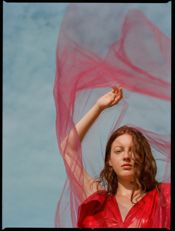 Red-haired woman in red dress with a pink tulle veil flowing against a blue sky in an outdoor fashion portrait, eye-level view.