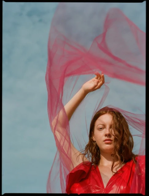 Red-haired woman in red dress with a pink tulle veil flowing against a blue sky in an outdoor fashion portrait, eye-level view.