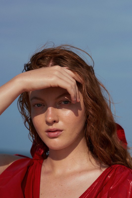 Close-up shot of a red-haired woman in a red dress with her hand shading her face against a clear blue sky, showcasing natural beauty.
