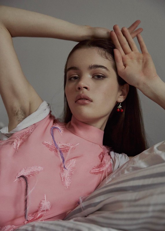 A young woman with fair skin, wearing a pink patterned top and cherry earrings, poses in a relaxed position on a bed with striped sheets.