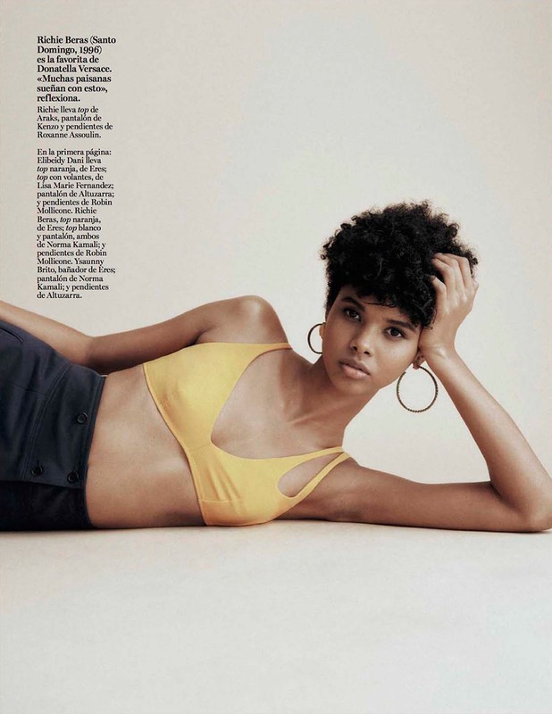A young woman with dark curly hair lies on a light surface, wearing a yellow top and navy bottoms, gazing directly at the camera.
