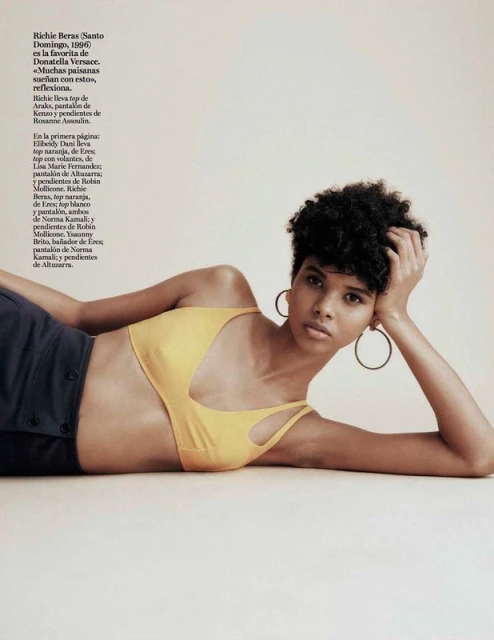 A young woman with dark curly hair lies on a light surface, wearing a yellow top and navy bottoms, gazing directly at the camera.