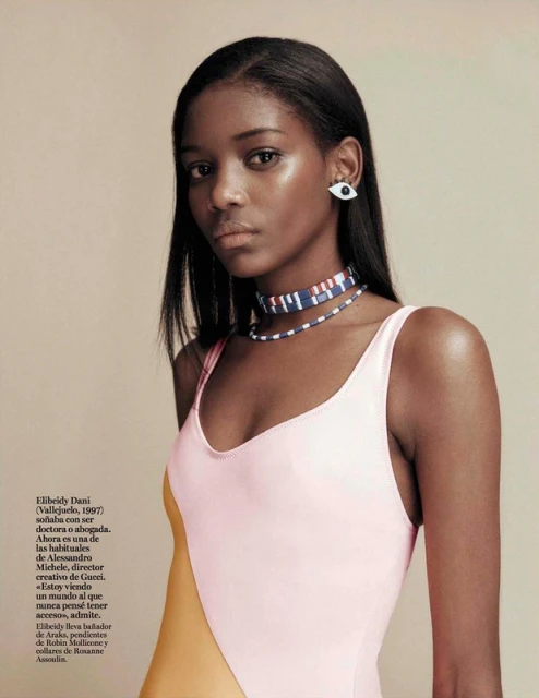 Close-up portrait of a Black woman with straight hair wearing an eye earring and necklace, a color-blocked one-piece swimsuit, against a tan backdrop.