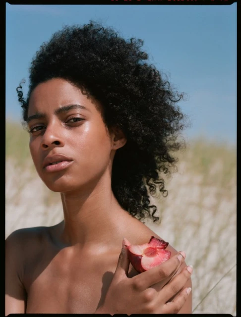 Close-up studio shot of a Black woman with curly hair holding a halved peach, against a background of sand dunes and a clear sky.