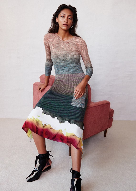 Elegant woman in a multicolored ombre knit dress seated on a pink chair, posing in a studio with ankle-tie heeled shoes for a fashion shot.