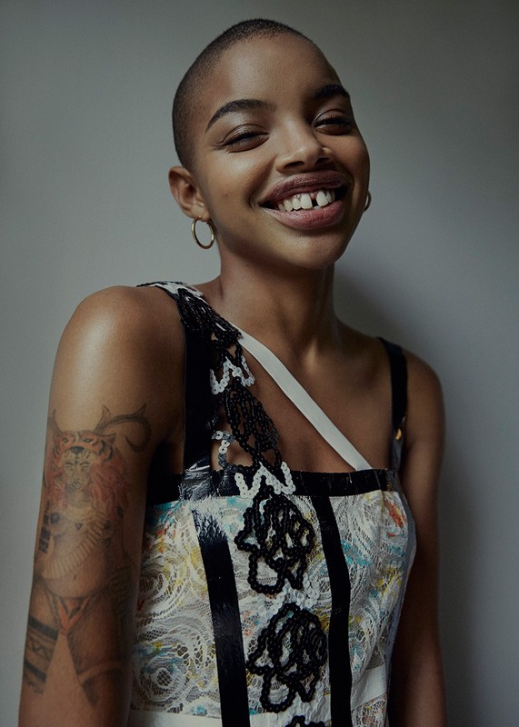 Smiling Black woman in patterned dress and gold hoop earrings poses against a gray wall in a studio shot; shoulder tattoo visible.
