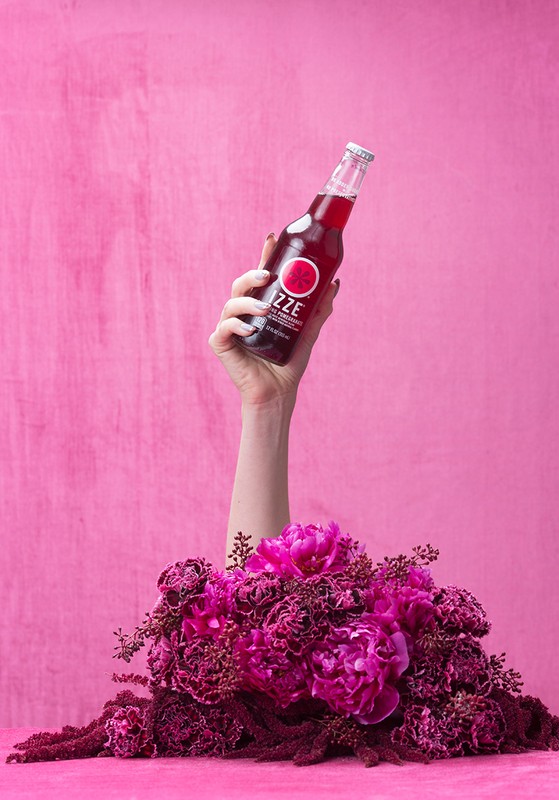 Arm holding a bottle of Izze surrounded by pink flowers in a studio setting against a pink background. Beverage product photography.