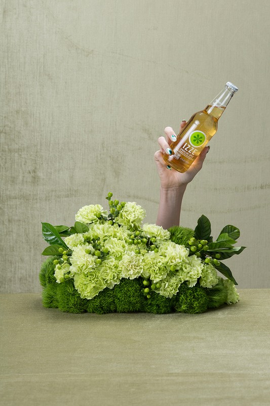 An arm raises a glass bottle of Izze sparkling juice from a bed of vibrant green carnations against a mottled green backdrop.