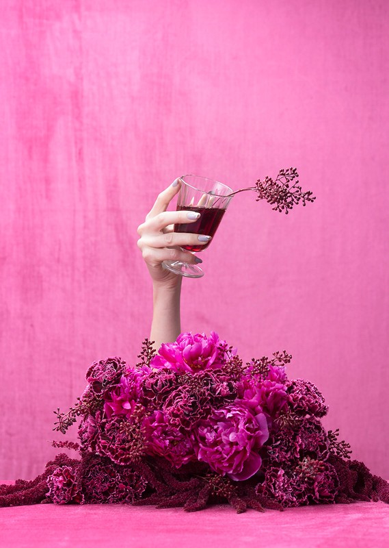 Hand holding glass with red liquid against pink background with a bouquet of flowers. Festive, celebratory.
