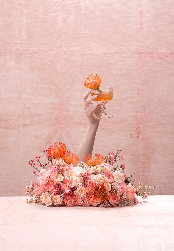 Hand holding cocktail glass with orange flower garnish above a floral arrangement against a textured pink backdrop. 