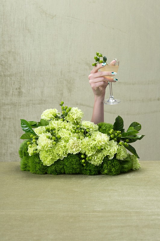 Elegant arm raises a champagne coupe amidst a green floral display with carnations and verdant foliage against a pale-green textured background.