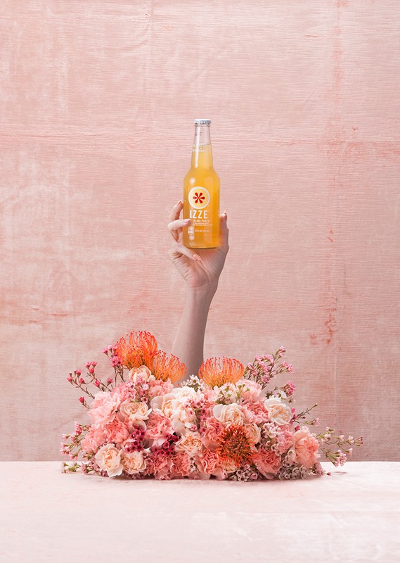 Hand holds an Izze sparkling juice drink above a pink floral arrangement against a matching pink textured background, studio shot.