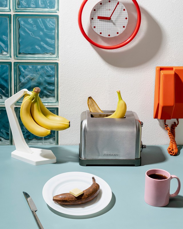 Still life of ripe and rotting bananas: a bunch on a stand, one in a toaster, one on a plate with butter, against an aqua and white wall.