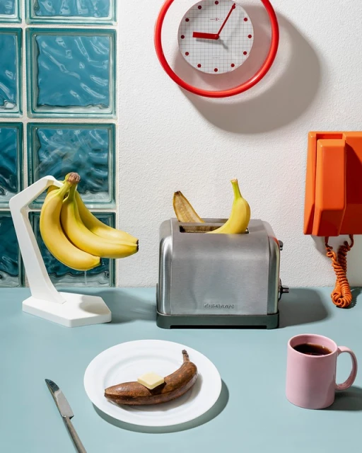 Still life of ripe and rotting bananas: a bunch on a stand, one in a toaster, one on a plate with butter, against an aqua and white wall.