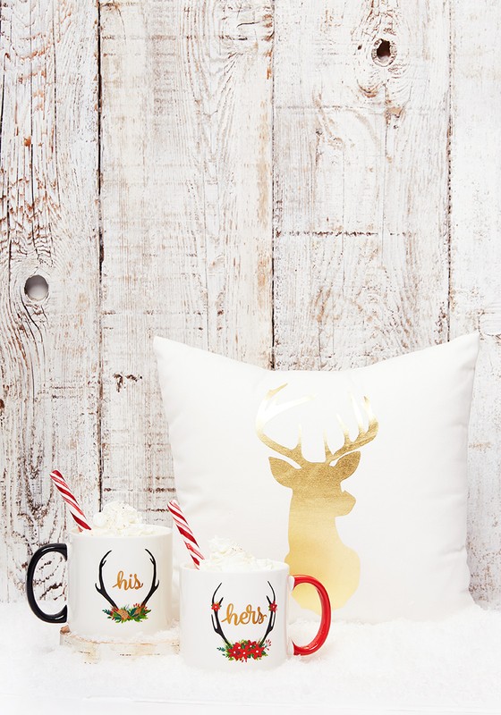 Festive mugs with 'his' and 'hers' text, marshmallow toppings, candy cane straws, and a gold reindeer pillow against a rustic wooden backdrop.