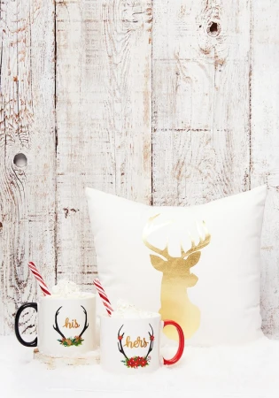 Festive mugs with 'his' and 'hers' text, marshmallow toppings, candy cane straws, and a gold reindeer pillow against a rustic wooden backdrop.