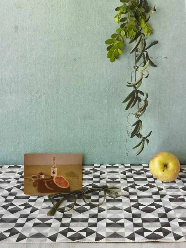 Still life featuring a painting, eyeglasses, an apple, and greenery against a textured, light blue wall and geometric tablecloth.