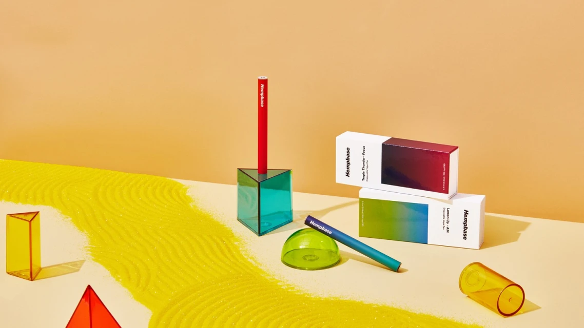 Still life of colorful geometric shapes, vape pens, and product boxes arranged on a tan surface with a yellow sand-like texture.