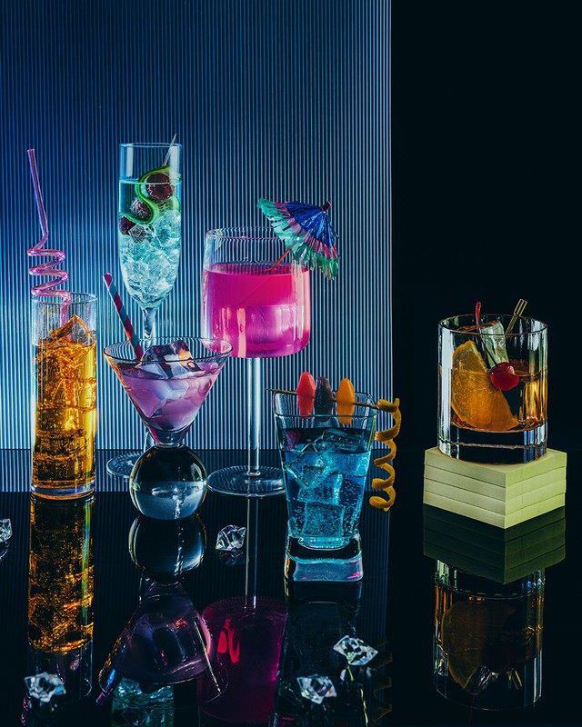 Arrangement of six colorful cocktails in various stemmed glasses, garnished with fruit and straws, on a glossy black surface.