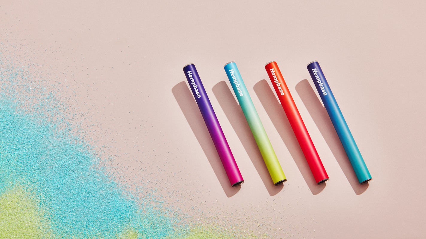 Four colorful Hempbase pens in purple, teal, red, and blue are scattered on a pink background with teal and green powder.