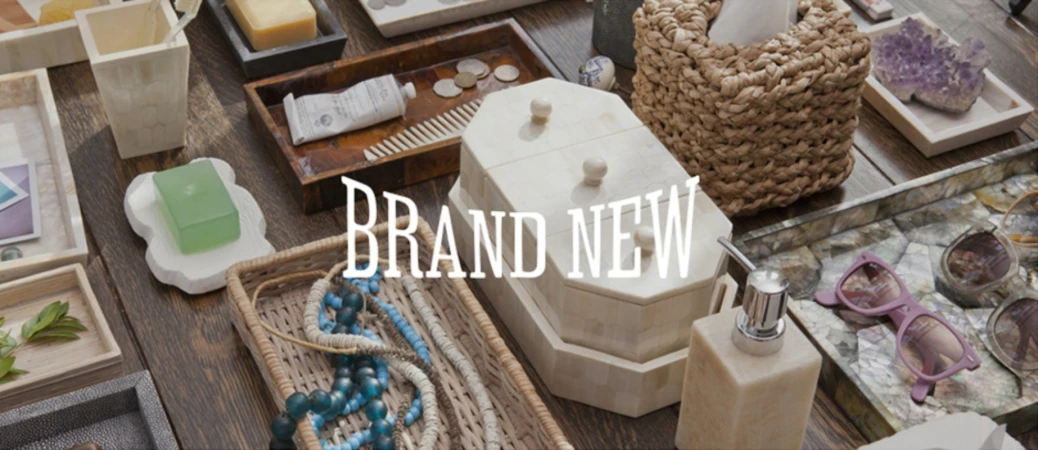 Various neutral-toned home goods arranged on a dark wood surface including bath accessories, jewelry, sunglasses, and decorative objects.