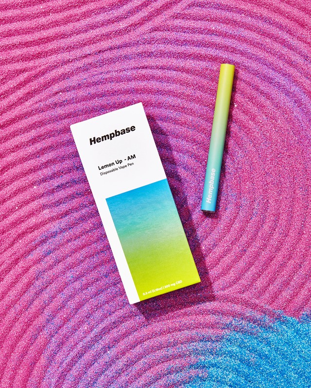 Product shot of Hempbase Lemon Up AM disposable vape pen and box on textured pink glitter surface with blue glitter accent.