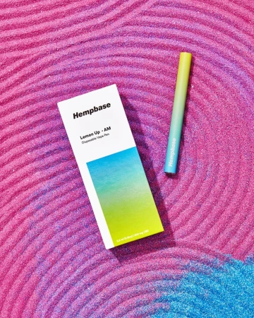 Product shot of Hempbase Lemon Up AM disposable vape pen and box on textured pink glitter surface with blue glitter accent.