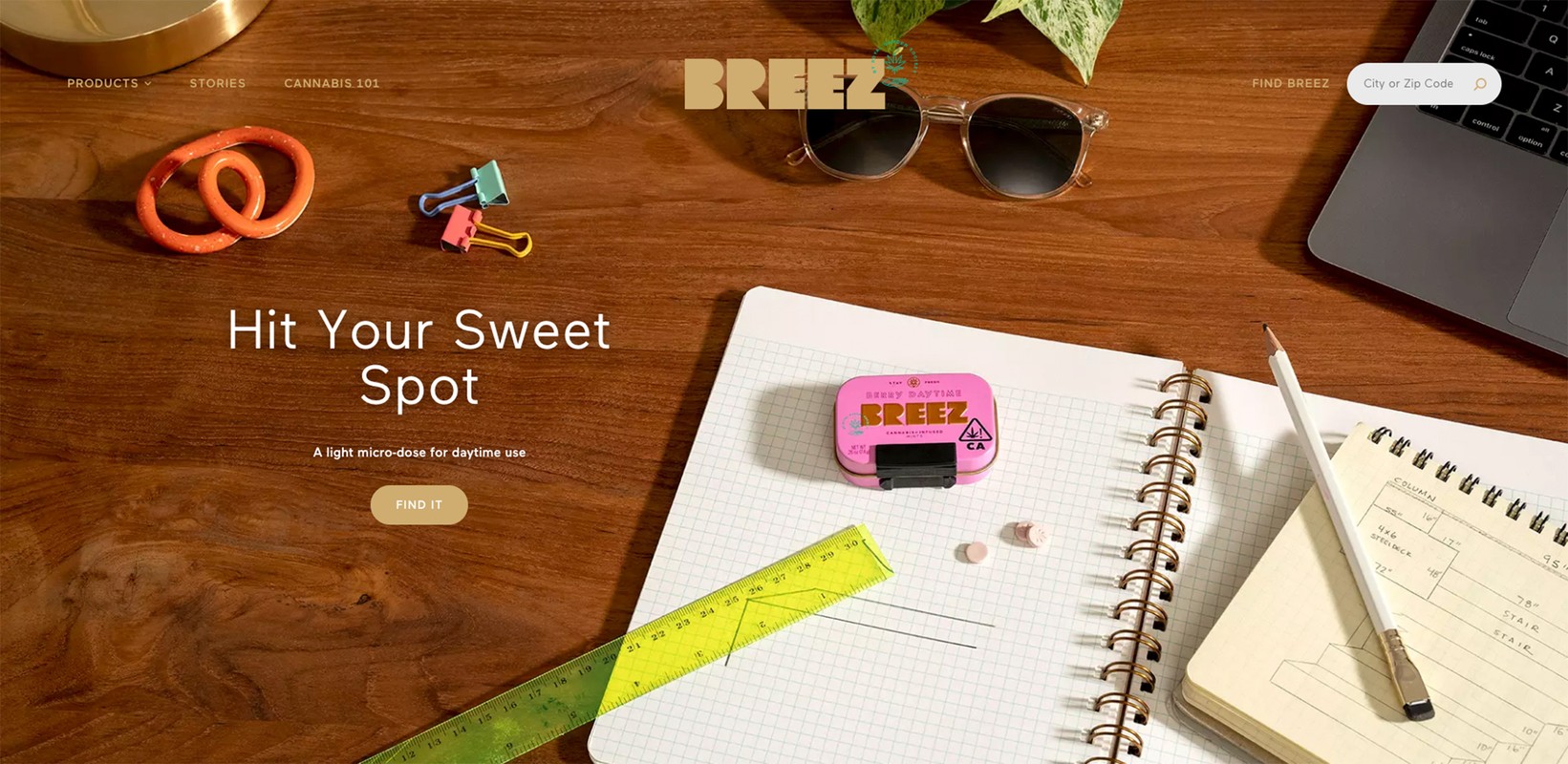 Overhead shot of a wood desk with a laptop, notebooks, sunglasses, and a pink tin of Breez cannabis products, conveying a modern, organized workspace.