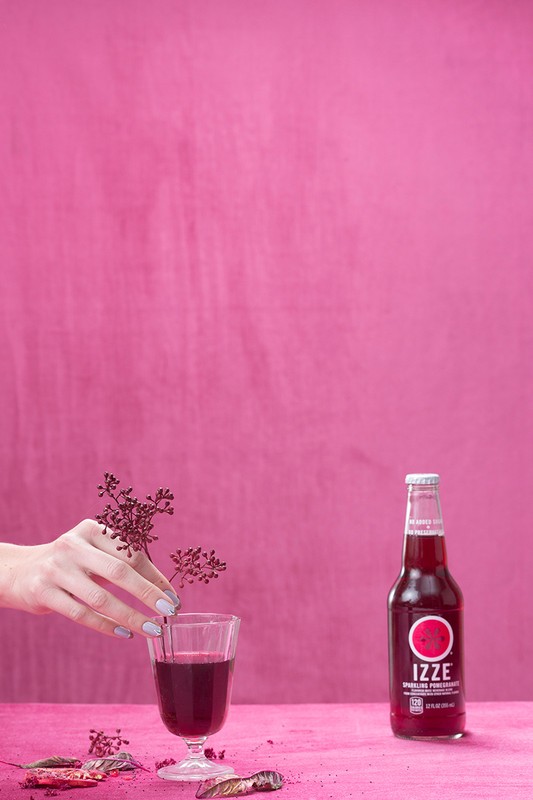 A bottle of Izze soda and a stemmed glass are staged in front of a pink background, with a hand placing a small branch in the glass.