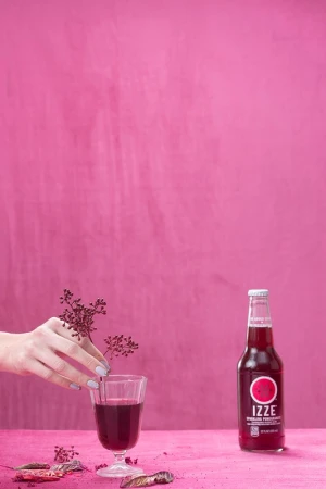 A bottle of Izze soda and a stemmed glass are staged in front of a pink background, with a hand placing a small branch in the glass.