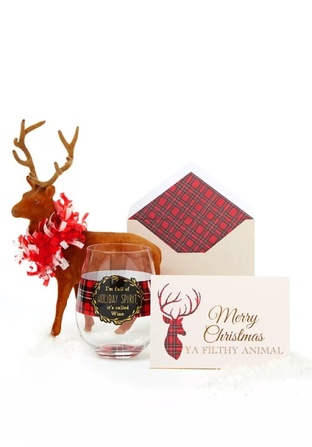 Holiday still life featuring a plush reindeer, wine glass with plaid accent, envelope, and greeting card on a white surface.