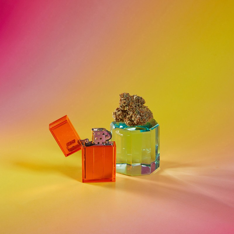 Still life photograph of an open orange lighter next to a glass container of marijuana buds on an orange and pink gradient background.