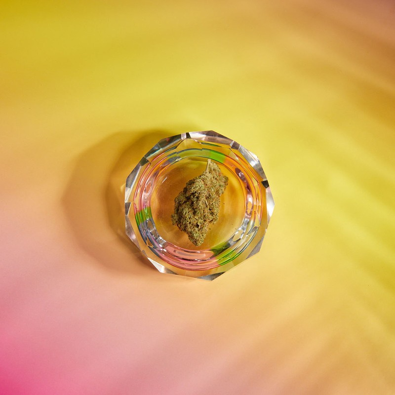 Overhead studio shot of a cannabis bud inside a faceted glass ashtray, isolated against an ombre yellow and pink background.