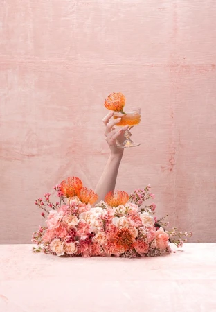 Hand holding a cocktail coupe garnished with a flower behind a dense pink floral arrangement against a textured pink background.