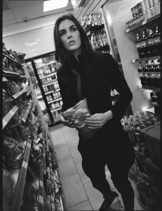 Monochrome photograph of an adult woman in casual clothing holding a bag of chips in a convenience store aisle.
