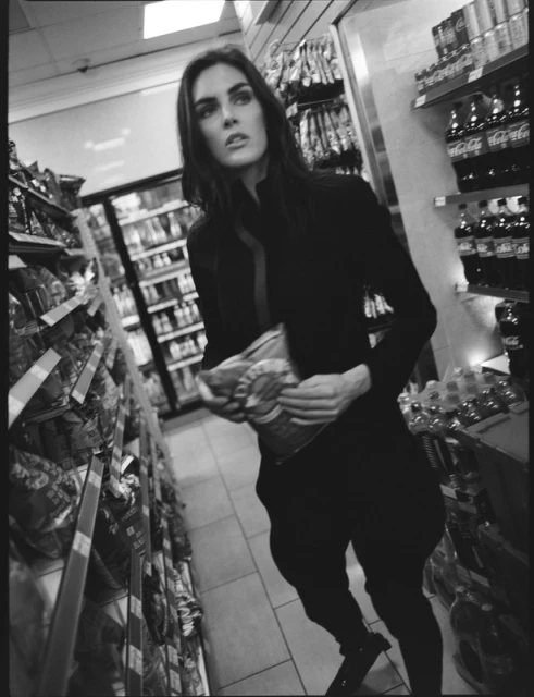 Monochrome photograph of an adult woman in casual clothing holding a bag of chips in a convenience store aisle.