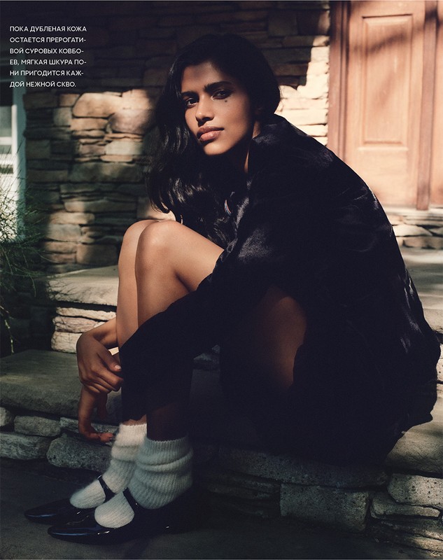 Young woman with dark hair and a dark jacket poses in black Mary Jane shoes and white socks, sitting on stone steps, looking at camera.