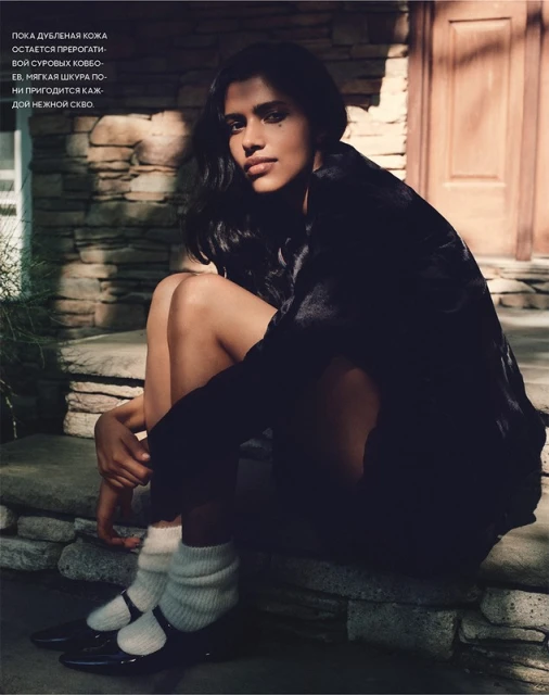 Young woman with dark hair and a dark jacket poses in black Mary Jane shoes and white socks, sitting on stone steps, looking at camera.