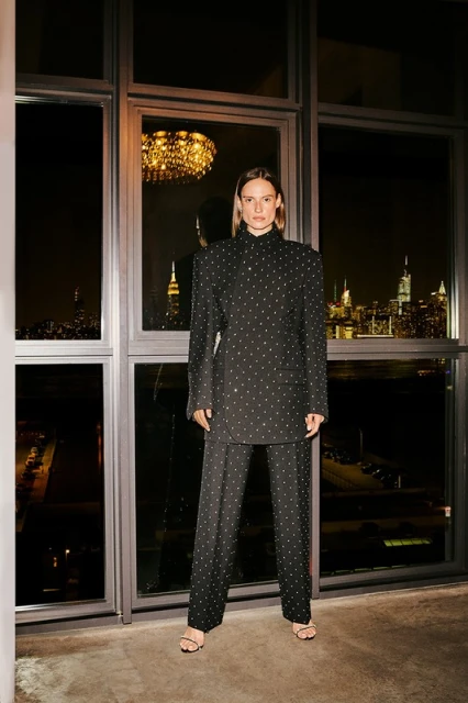 Woman in a studded black blazer and pants stands in front of a large window with a cityscape view at night. Fashion shot, indoor.