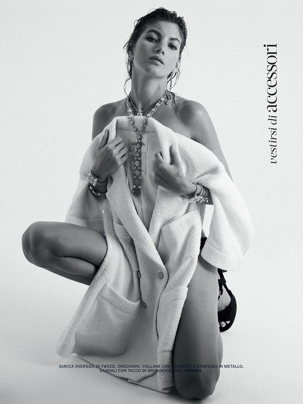 Monochrome studio shot of a woman kneeling, adorned with a necklace and bracelets, draped in a tweed jacket, against a white backdrop.