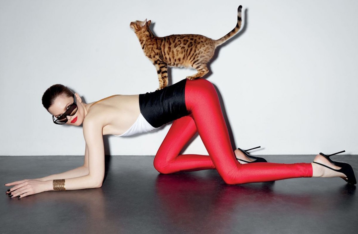 Fashion shot of a woman in a bodysuit and leggings posing with a cat on her back in a bright studio with black flooring.