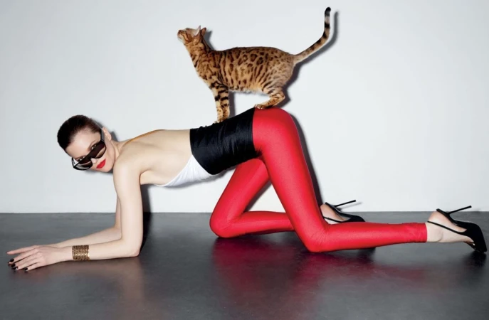 Fashion shot of a woman in a bodysuit and leggings posing with a cat on her back in a bright studio with black flooring.