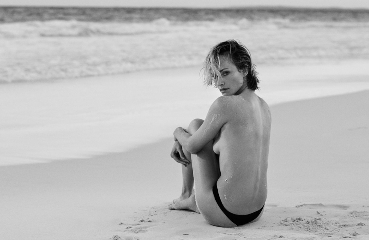 Black and white portrait of a woman sitting on a sandy beach by the ocean with crashing waves in the background, looking back.