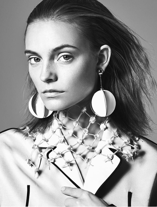 Striking black and white fashion portrait of a woman with bold, round earrings and a distinctive geometric necklace against a neutral backdrop.