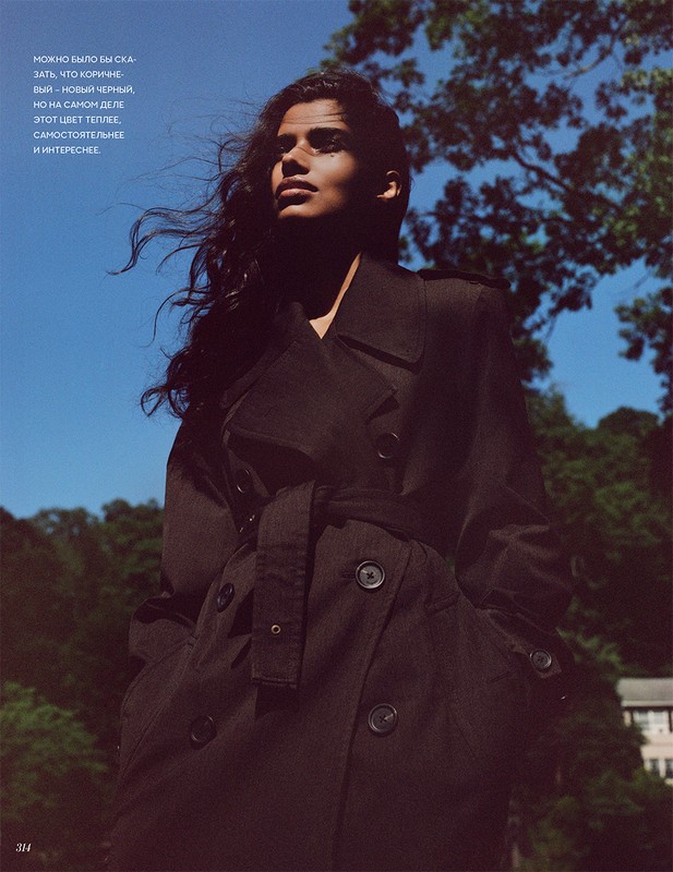 A young adult woman models a trench coat in an outdoor shot under a clear blue sky with scattered trees in the background.