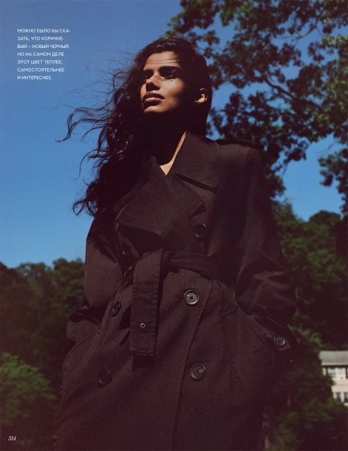 A young adult woman models a trench coat in an outdoor shot under a clear blue sky with scattered trees in the background.
