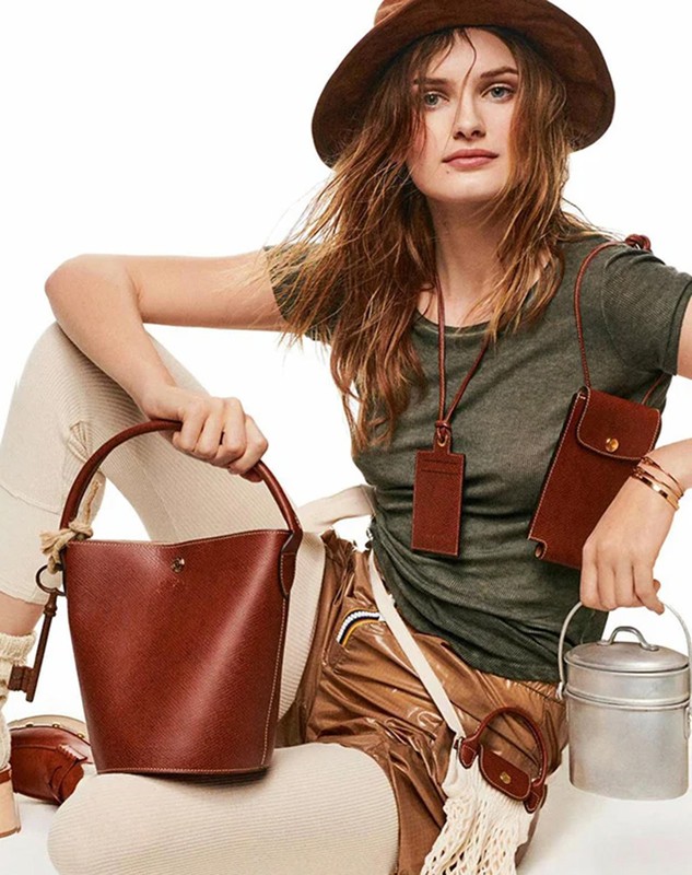 A woman in a hat sits surrounded by several handbags and fashion accessories in a light studio setting, showcasing brown and tan tones.