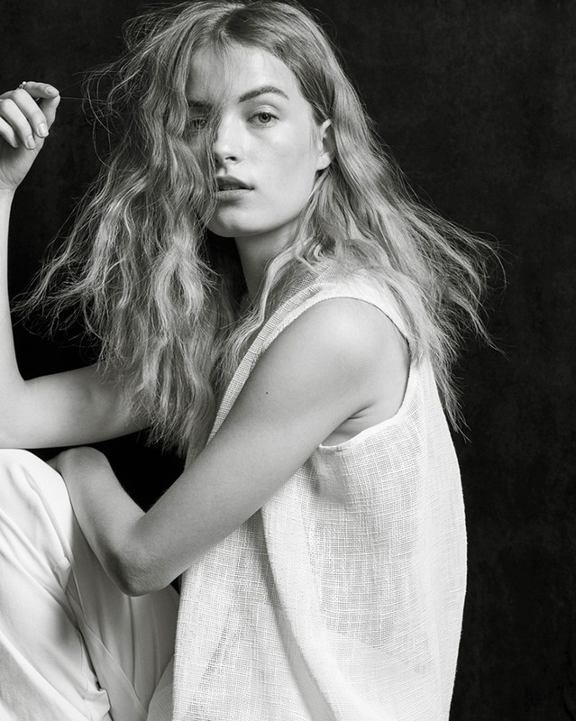 Portrait of a woman with tousled long hair wearing a white sleeveless top against a dark textured background.